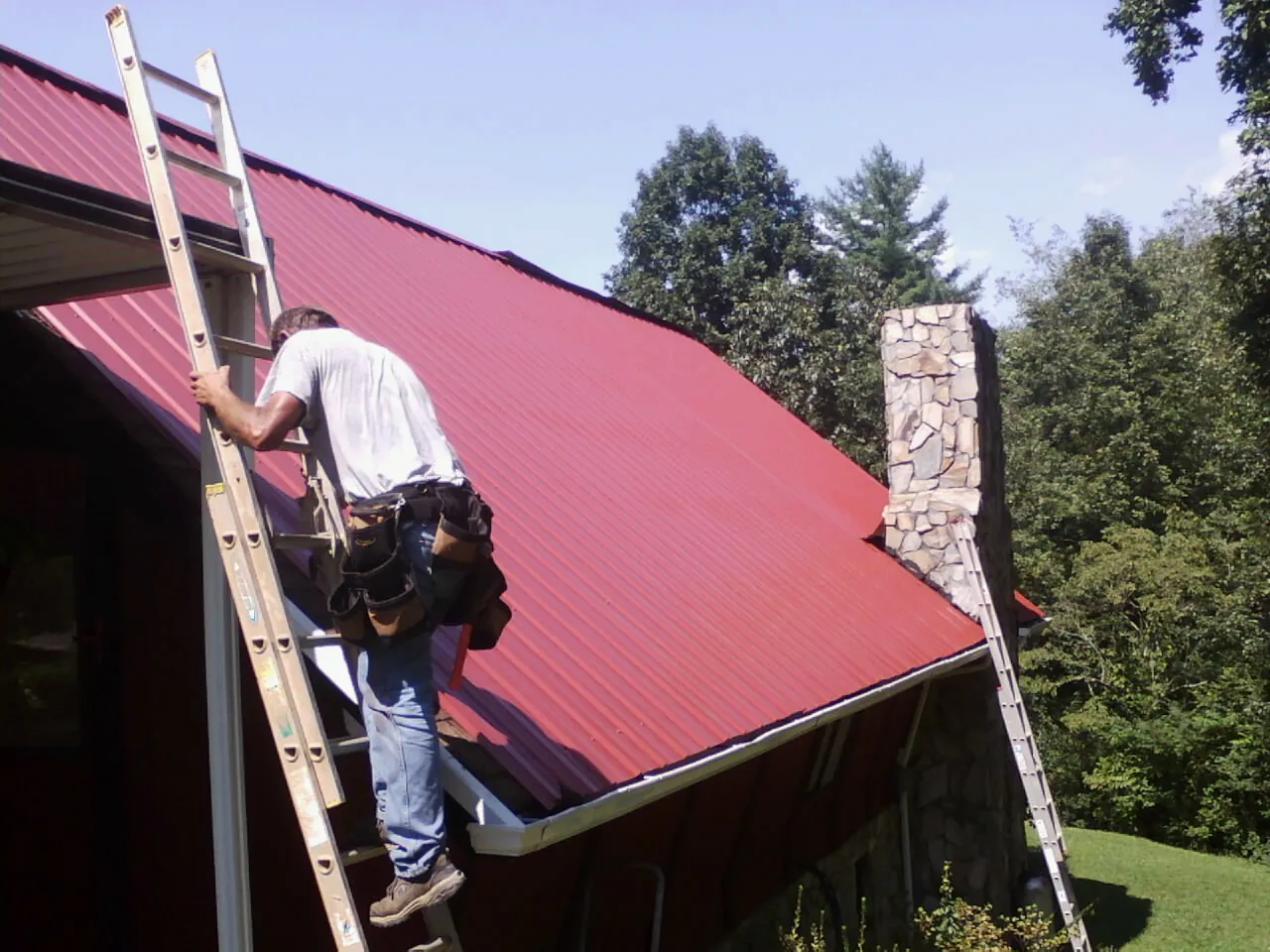 Roofer on a ladder beside a red metal roof and stone chimney — Soffit & Fascia Repair by Roofing Brothers in Woodridge